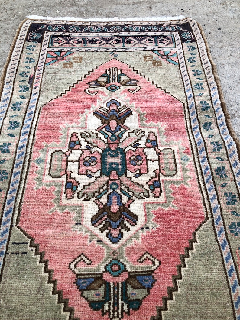 May include: A vintage pink and green rug with a traditional floral pattern. The rug has a fringed edge and is made of wool.