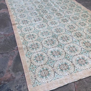 May include: A vintage rug with a cream background and a repeating pattern of green and brown floral motifs. The rug has a fringed edge.