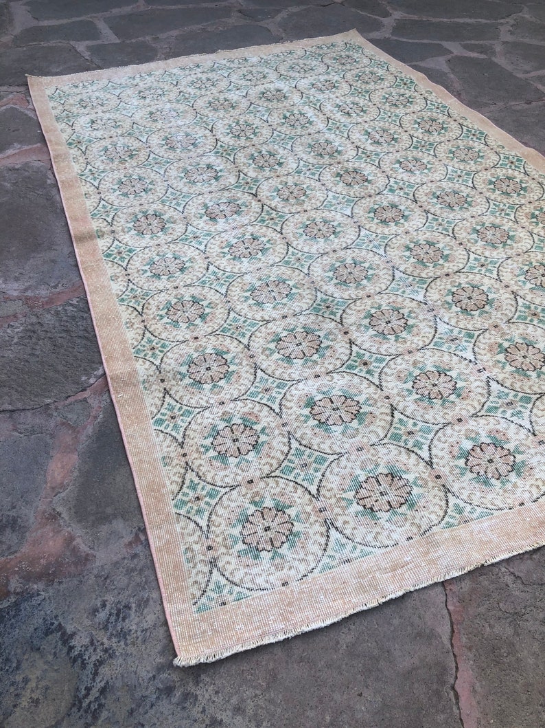 May include: A vintage rug with a cream background and a repeating pattern of green and brown floral motifs. The rug has a fringed edge.