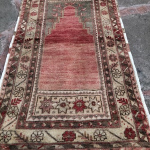 May include: A vintage Turkish prayer rug with a red center and a beige border with intricate floral patterns. The rug has a fringe on all sides.