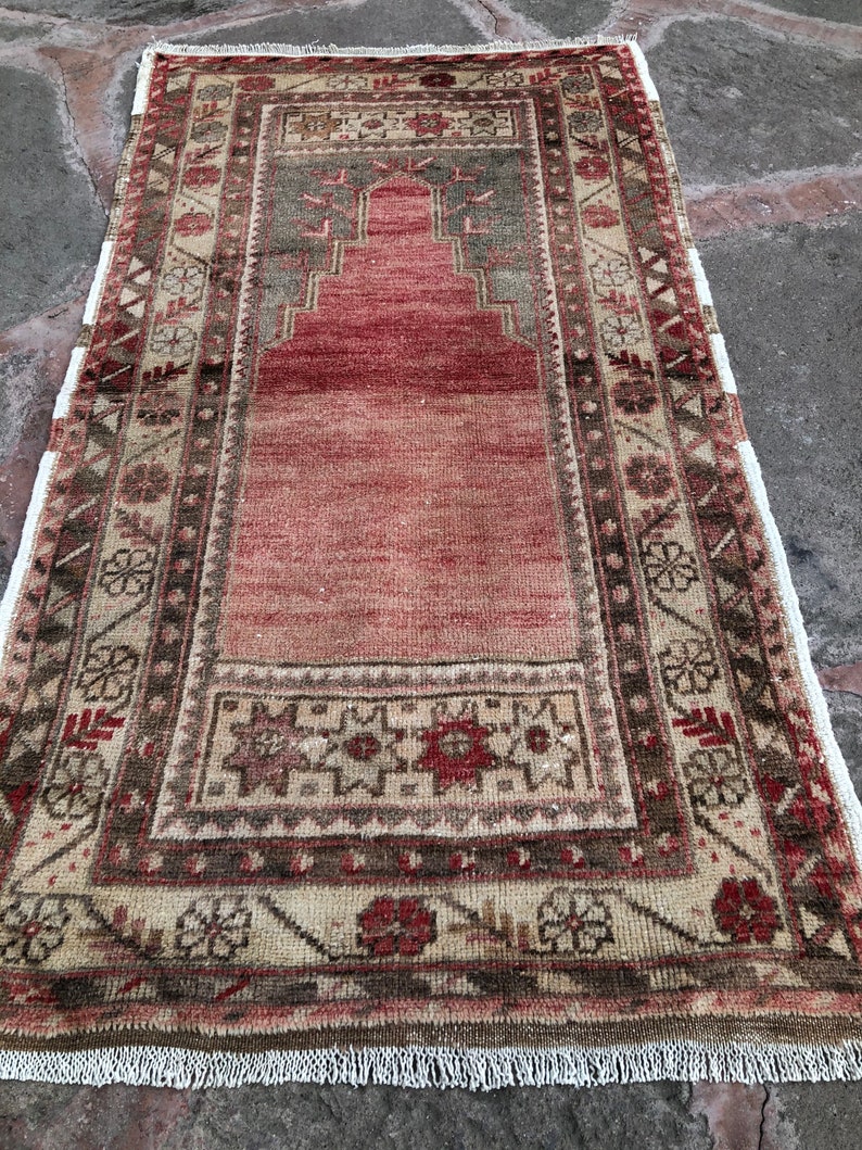 May include: A vintage Turkish prayer rug with a red center and a beige border with intricate floral patterns. The rug has a fringe on all sides.
