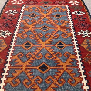 May include: A rectangular, hand-woven rug with a central blue field featuring orange geometric patterns. The rug has a red border with white and dark blue accents. The top edge has a brown fringe.