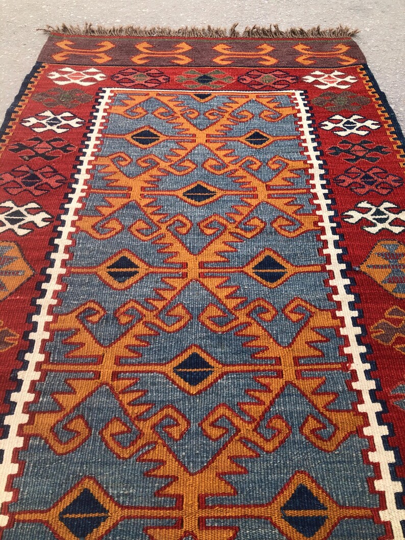 May include: A rectangular, hand-woven rug with a central blue field featuring orange geometric patterns. The rug has a red border with white and dark blue accents. The top edge has a brown fringe.
