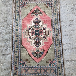 May include: A vintage rug with a pink, green, and blue geometric pattern. The rug has a fringe border and is made of wool.