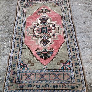May include: A vintage Turkish rug with a pink and green geometric pattern. The rug has a fringed edge and is made of wool.