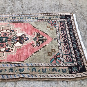 May include: A vintage rug with a pink, green, and brown geometric pattern. The rug has a fringe border and is made of wool.