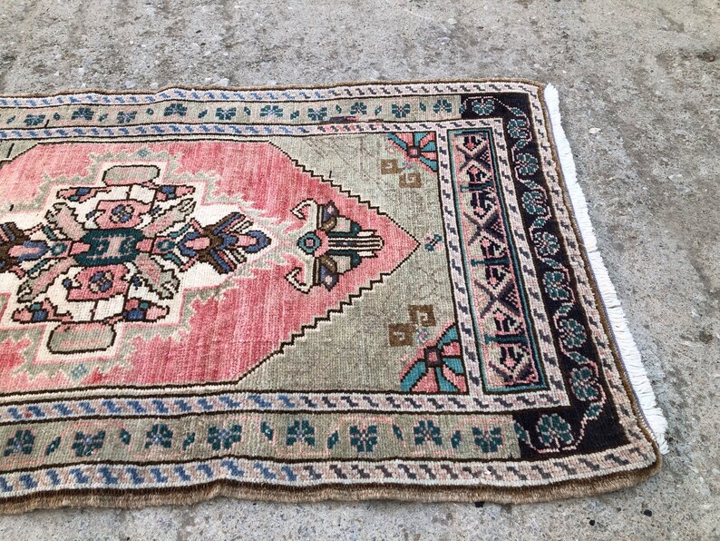 May include: A vintage rug with a pink, green, and brown geometric pattern. The rug has a fringe border and is made of wool.