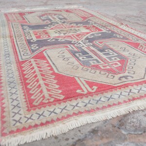 May include: A vintage Turkish rug with a red, beige, and blue geometric pattern. The rug has a fringe border and is made of wool.