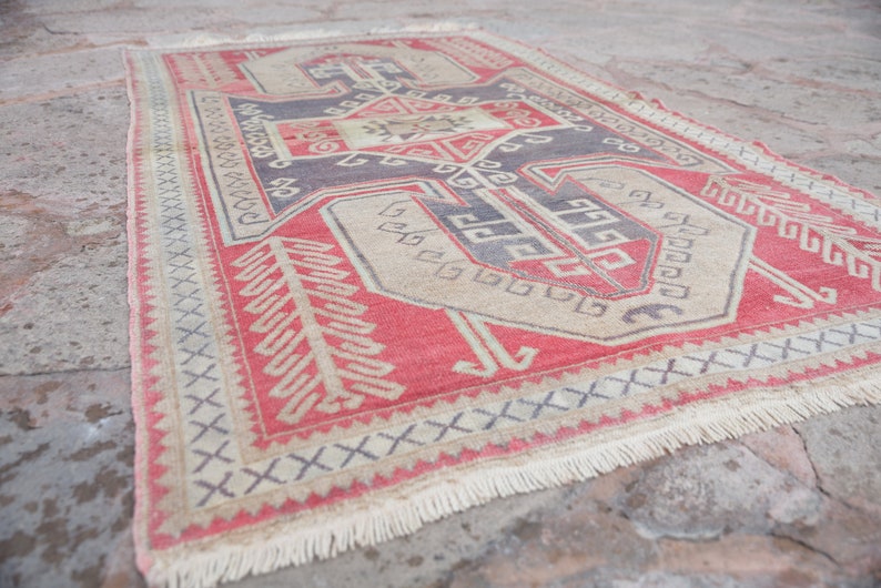 May include: A vintage Turkish rug with a red, beige, and blue geometric pattern. The rug has a fringe border and is made of wool.