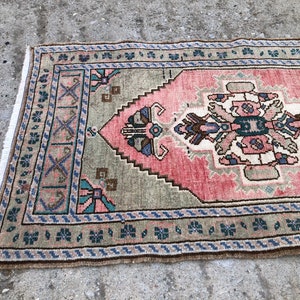 May include: A vintage rug with a pink and green geometric pattern. The rug has a fringed edge and is made of wool.