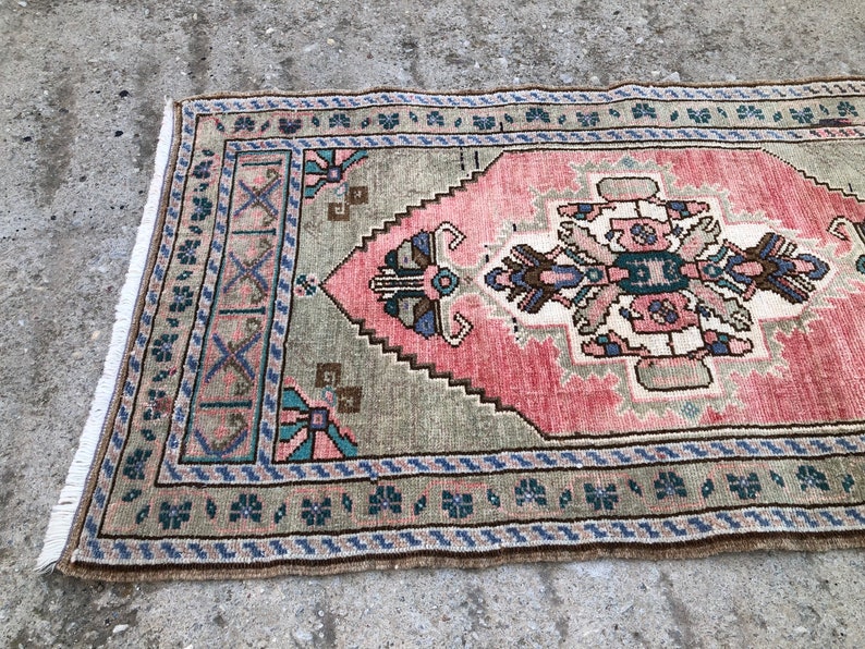 May include: A vintage rug with a pink and green geometric pattern. The rug has a fringed edge and is made of wool.