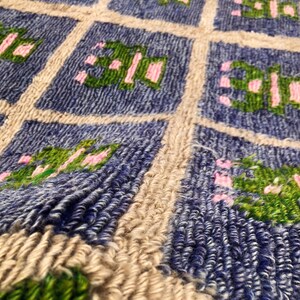 以下が含まれることがあります： 青い正方形の繰り返し模様の、質感のあるラグのクローズアップ。各正方形には、緑とピンクの花柄のデザインが含まれています。ラグはベージュの縁取りがあり、厚手の織り素材で作られています。