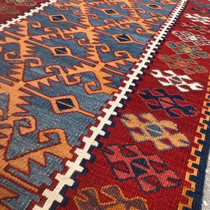 May include: A rectangular, hand-woven rug with a geometric pattern. The rug features a central design in shades of blue and orange, bordered by a white zigzag pattern and a red background. The rug's design includes various shapes and patterns.