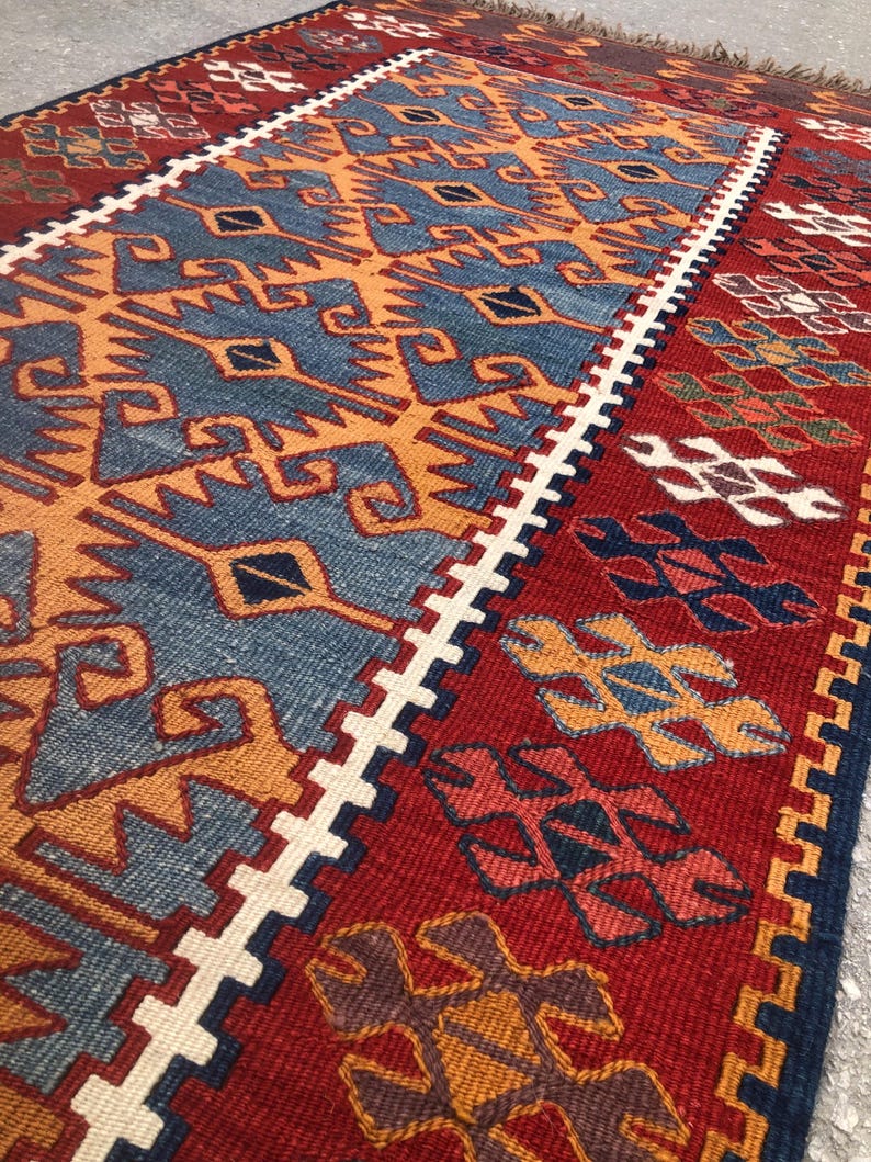 May include: A rectangular, hand-woven rug with a geometric pattern. The rug features a central design in shades of blue and orange, bordered by a white zigzag pattern and a red background. The rug's design includes various shapes and patterns.