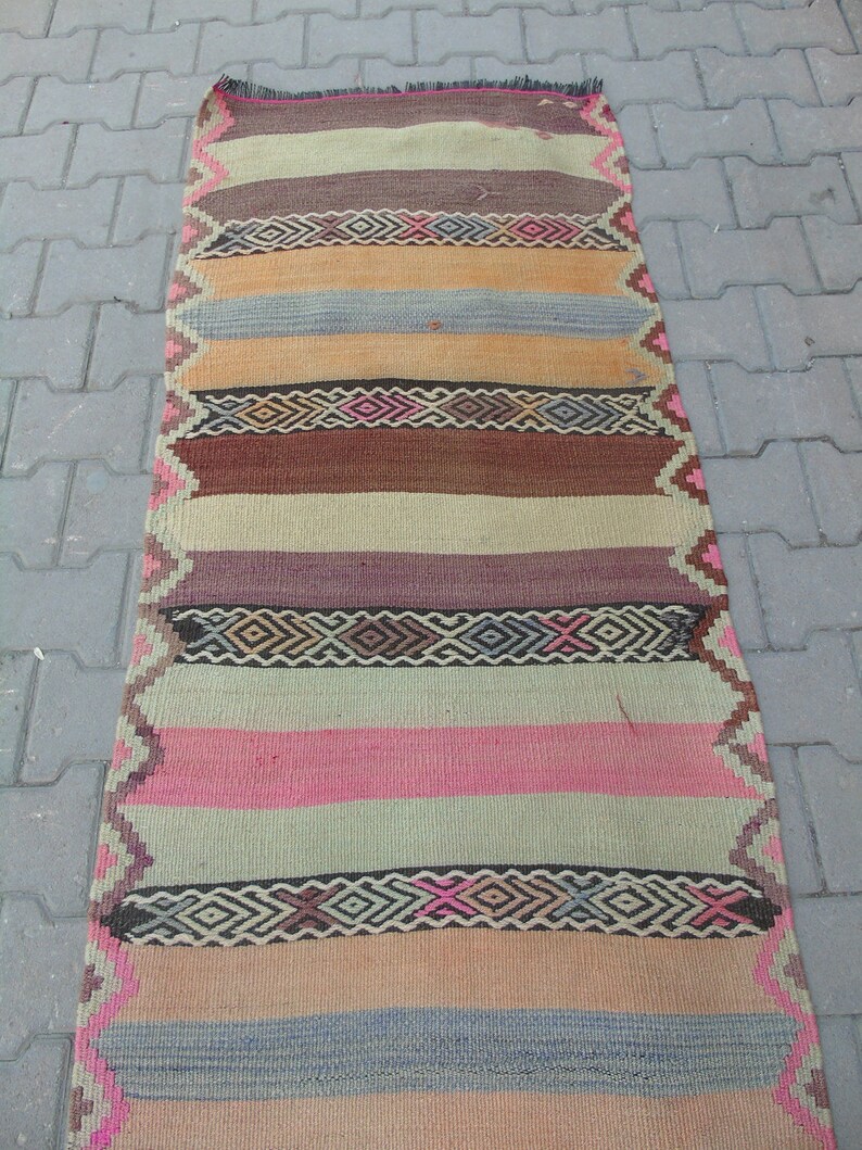 May include: A long, narrow, multi-colored rug with a striped pattern. The rug is made of woven wool and features a variety of colors, including brown, pink, beige, and blue.