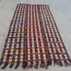 May include: A colorful, handwoven rug with a checkered pattern in shades of red, white, black, and orange. The rug has a fringe border and is made of wool.