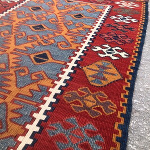 May include: A vibrant, hand-woven rug with a geometric design. The rug features a red background with intricate patterns in shades of blue, orange, and white. The edges are finished with a dark blue border and fringe.