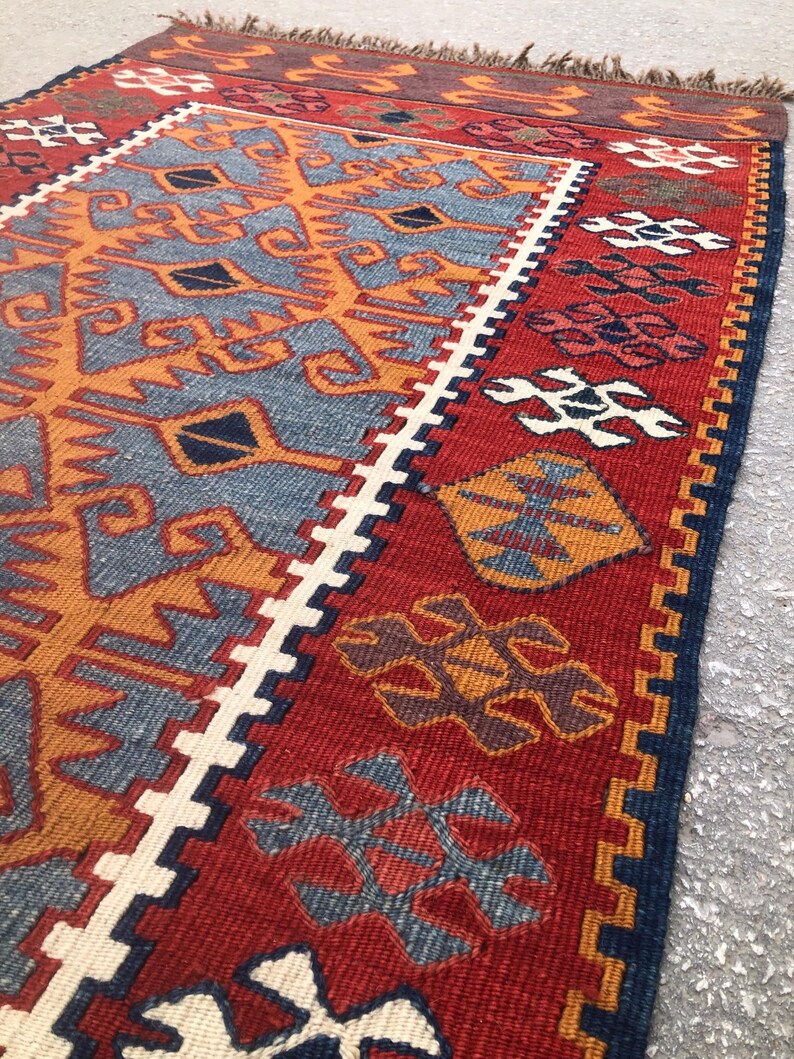 May include: A vibrant, hand-woven rug with a geometric design. The rug features a red background with intricate patterns in shades of blue, orange, and white. The edges are finished with a dark blue border and fringe.