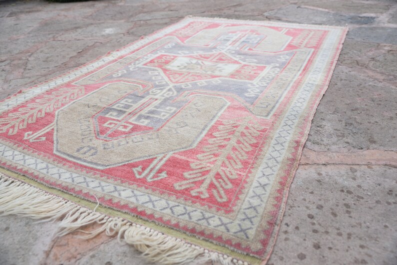 May include: A vintage Turkish rug with a red, beige, and blue geometric pattern. The rug has a fringe border and is lying on a concrete surface.