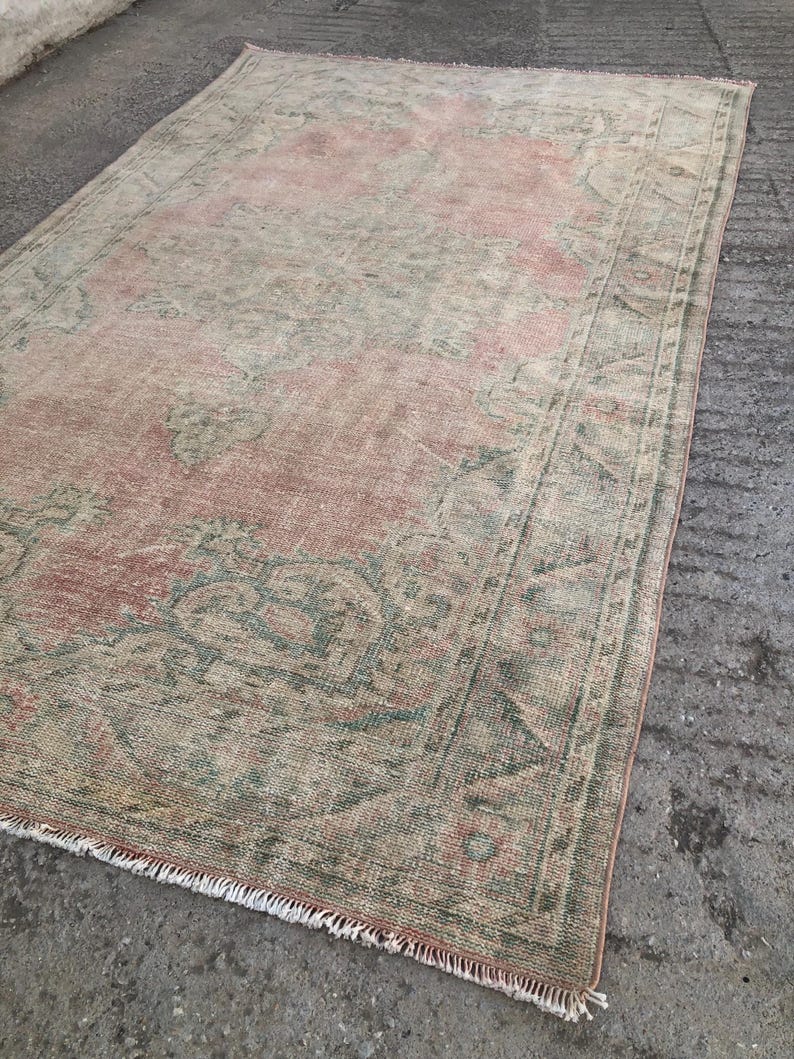 May include: A vintage rug with a faded pink and green floral pattern. The rug has a fringed edge and is made of wool.