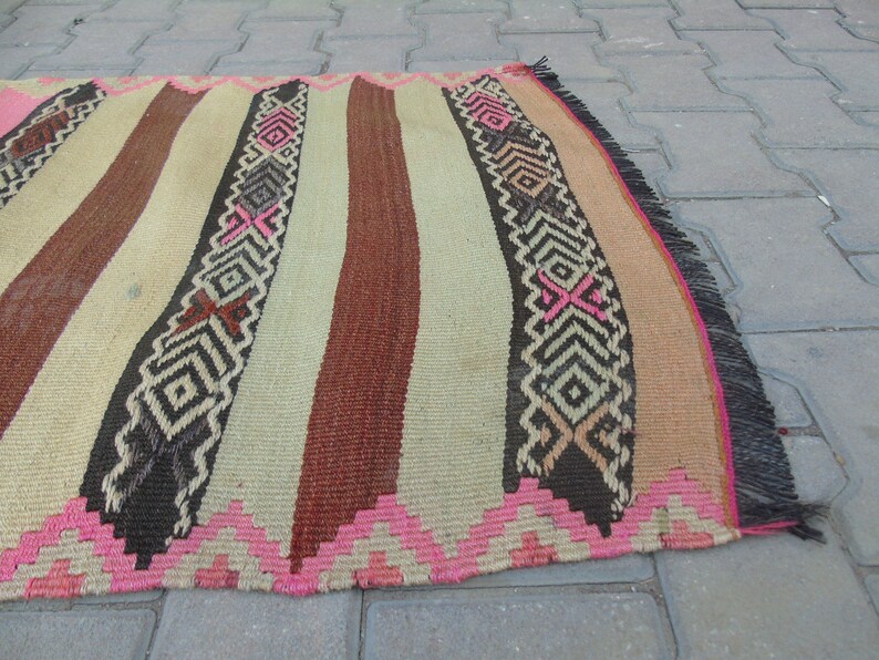 May include: A handwoven rug with a striped pattern in shades of brown, beige, and pink. The rug has a fringe border and intricate geometric designs.