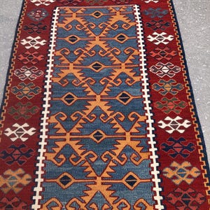 May include: A rectangular rug with a central blue field featuring orange geometric patterns. The rug has a red border with repeating patterns in white, orange, and blue. The top edge has a brown fringe.