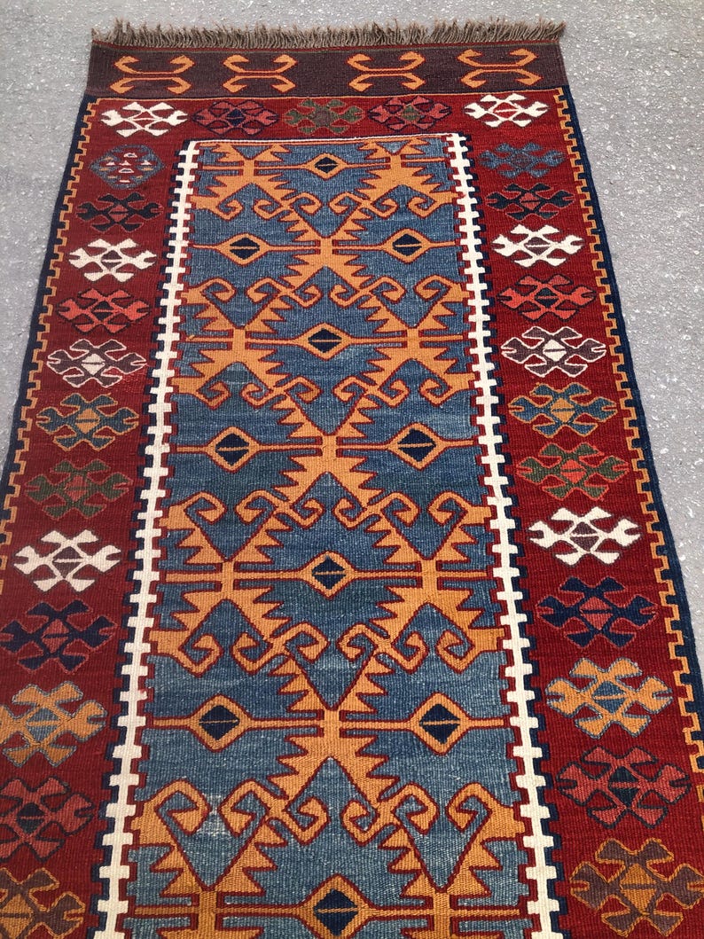 May include: A rectangular rug with a central blue field featuring orange geometric patterns. The rug has a red border with repeating patterns in white, orange, and blue. The top edge has a brown fringe.