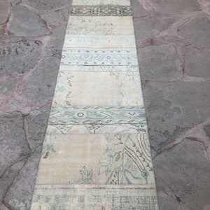 May include: A long, narrow, off-white rug with a faded, vintage look. The rug has a patchwork design with different patterns and colors, including green, brown, and pink. The rug is made of wool and has a soft, comfortable texture.