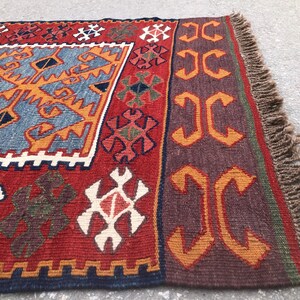 May include: A rectangular, hand-woven rug with a vibrant design. The rug features a red background with geometric patterns in orange, blue, white, and brown. The edges are finished with a fringe.