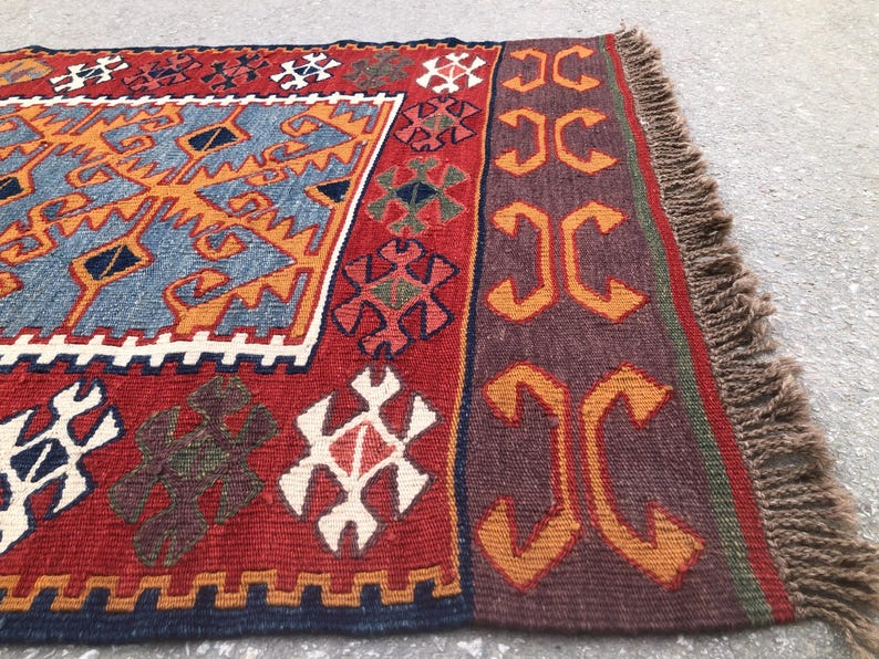 May include: A rectangular, hand-woven rug with a vibrant design. The rug features a red background with geometric patterns in orange, blue, white, and brown. The edges are finished with a fringe.