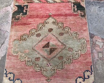 May include: A rectangular, vintage rug with a faded, distressed appearance. The rug features a central diamond-shaped design in muted green and brown, set against a pinkish-red background. Decorative borders and accents in cream, black, and blue complete the design.