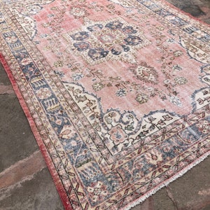 May include: A vintage pink and cream rug with a floral pattern and a blue and cream border. The rug has a fringed edge.