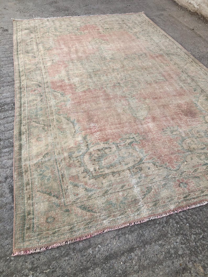 May include: A vintage rug with a faded pink and green floral pattern. The rug has a fringed edge and is made of a soft, woven material.