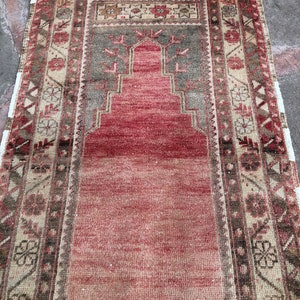 May include: A vintage Turkish prayer rug with a red center and a geometric pattern in shades of brown, beige, and green. The rug has a fringed edge.