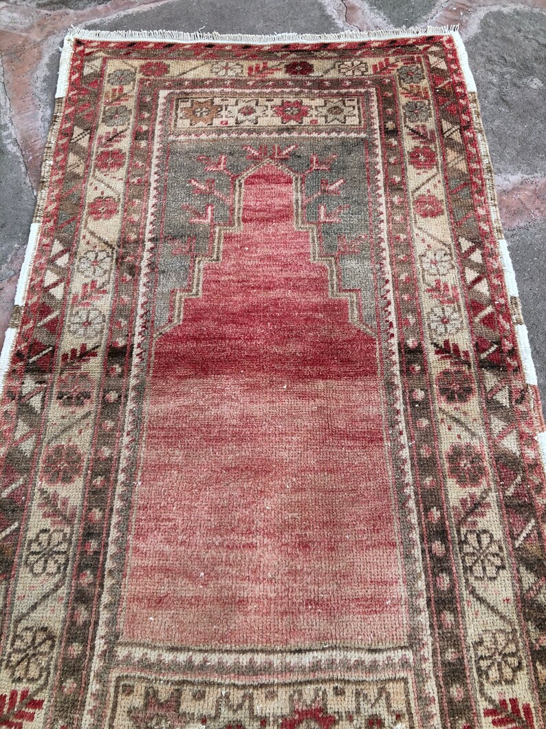 May include: A vintage Turkish prayer rug with a red center and a geometric pattern in shades of brown, beige, and green. The rug has a fringed edge.