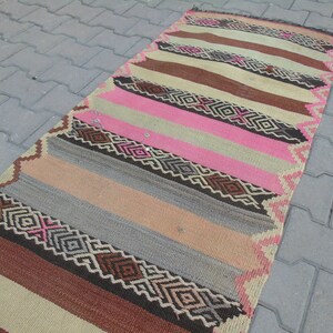May include: A long, narrow rug with a striped pattern in shades of brown, pink, beige, and gray. The rug has a geometric design with diamond shapes and intricate details.