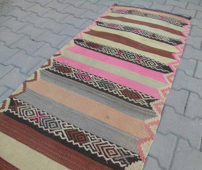 May include: A long, narrow rug with a striped pattern in shades of brown, pink, beige, and gray. The rug has a geometric design with diamond shapes and intricate details.