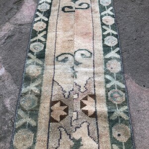 May include: A long, narrow rug with a cream background and green, brown, and white geometric patterns. The rug has a fringe on both ends.