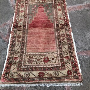 May include: A vintage Turkish prayer rug with a traditional design. The rug is beige with red, brown, and green accents. It has a fringed edge.