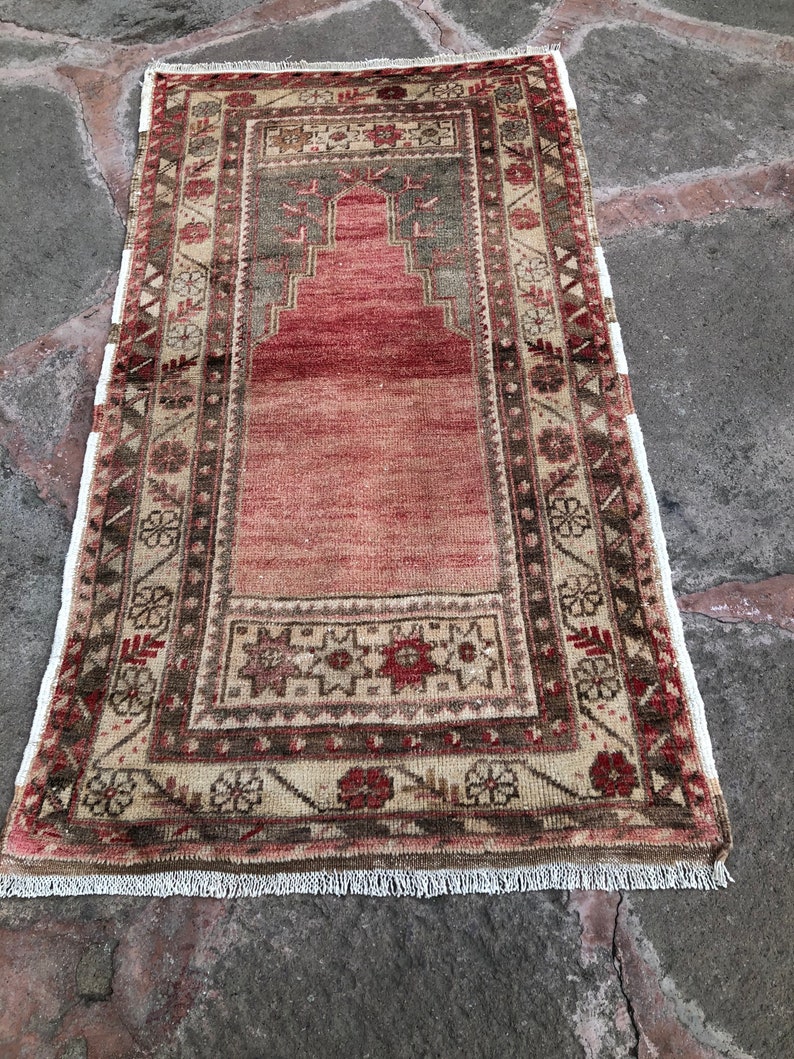 May include: A vintage Turkish prayer rug with a traditional design. The rug is beige with red, brown, and green accents. It has a fringed edge.