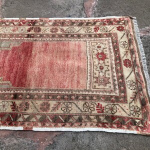 May include: A vintage Turkish rug with a red and cream color scheme. The rug has a geometric pattern with floral motifs and a fringe border.
