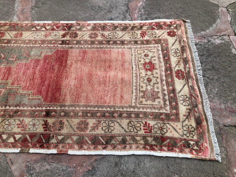 May include: A vintage Turkish rug with a red and cream color scheme. The rug has a geometric pattern with floral motifs and a fringe border.