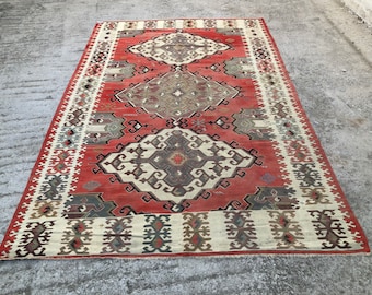 May include: A rectangular, hand-woven rug with a central geometric design in shades of red, cream, and brown. The rug features a repeating pattern along the borders and within the central field. The rug's dimensions are not visible in the image.