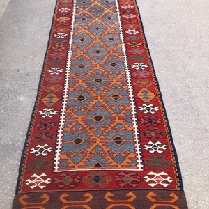 May include: A rectangular, patterned rug with a central design in blue and orange, framed by a red border. The rug features geometric patterns and a fringed edge, suitable for home decor.