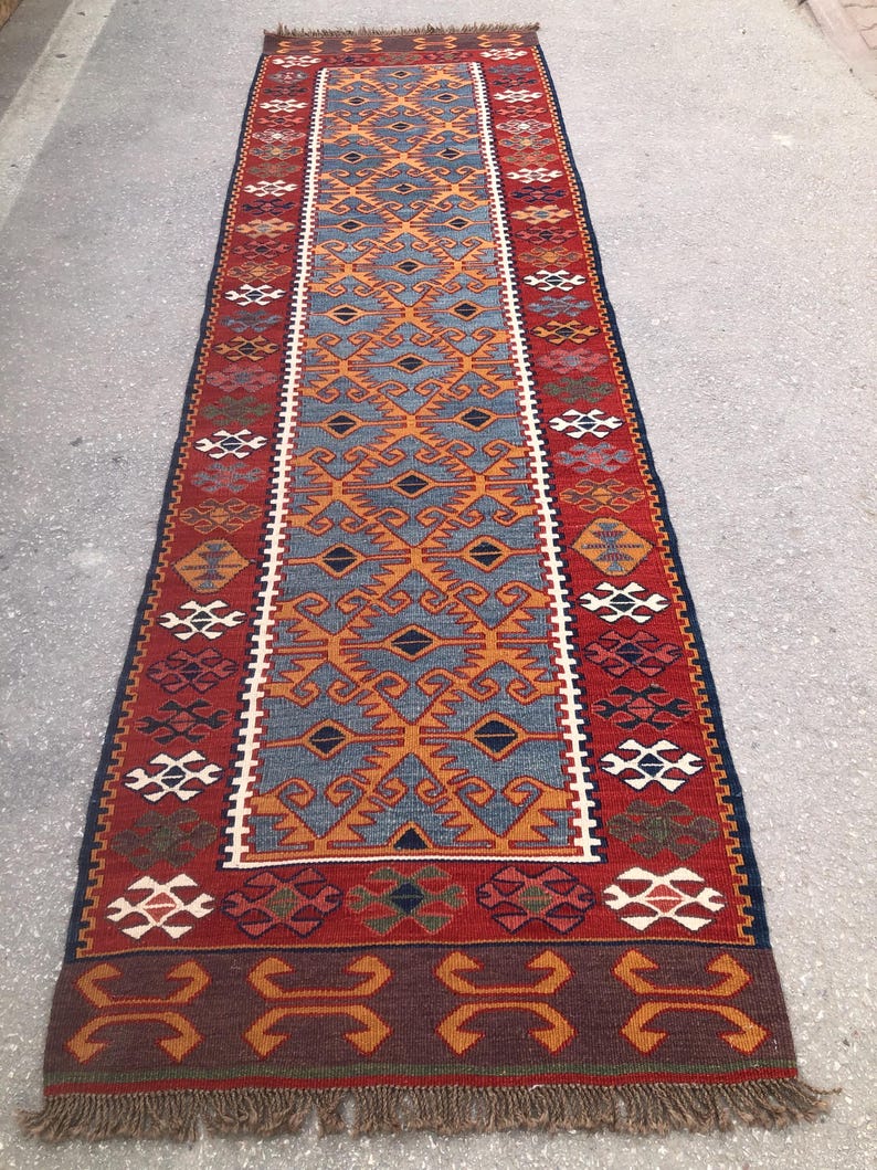 May include: A rectangular, patterned rug with a central design in blue and orange, framed by a red border. The rug features geometric patterns and a fringed edge, suitable for home decor.