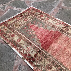 May include: A vintage Turkish prayer rug with a red, beige, and brown geometric pattern. The rug has a white fringe border.