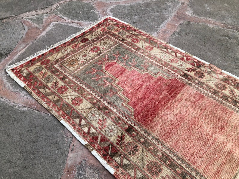 May include: A vintage Turkish prayer rug with a red, beige, and brown geometric pattern. The rug has a white fringe border.