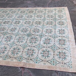 May include: A vintage rug with a cream background and a repeating pattern of green and brown floral motifs. The rug has a light pink border.
