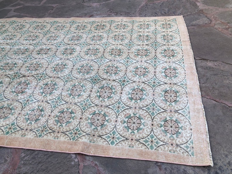 May include: A vintage rug with a cream background and a repeating pattern of green and brown floral motifs. The rug has a light pink border.
