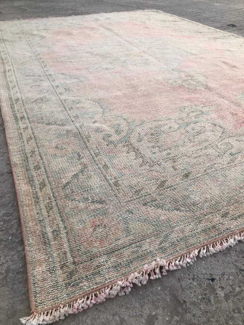May include: A vintage rug with a faded pink and green floral pattern. The rug has a fringed edge and is made of a soft, woven material.